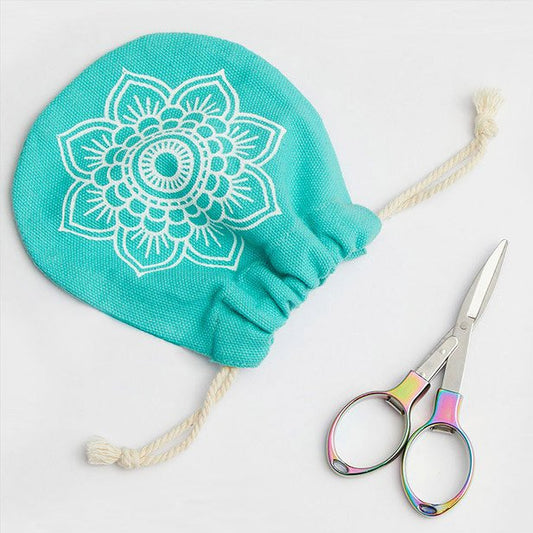 A round turquoise drawstring pouch with a white floral pattern next to a pair of iridescent color scissors on a light grey background.
