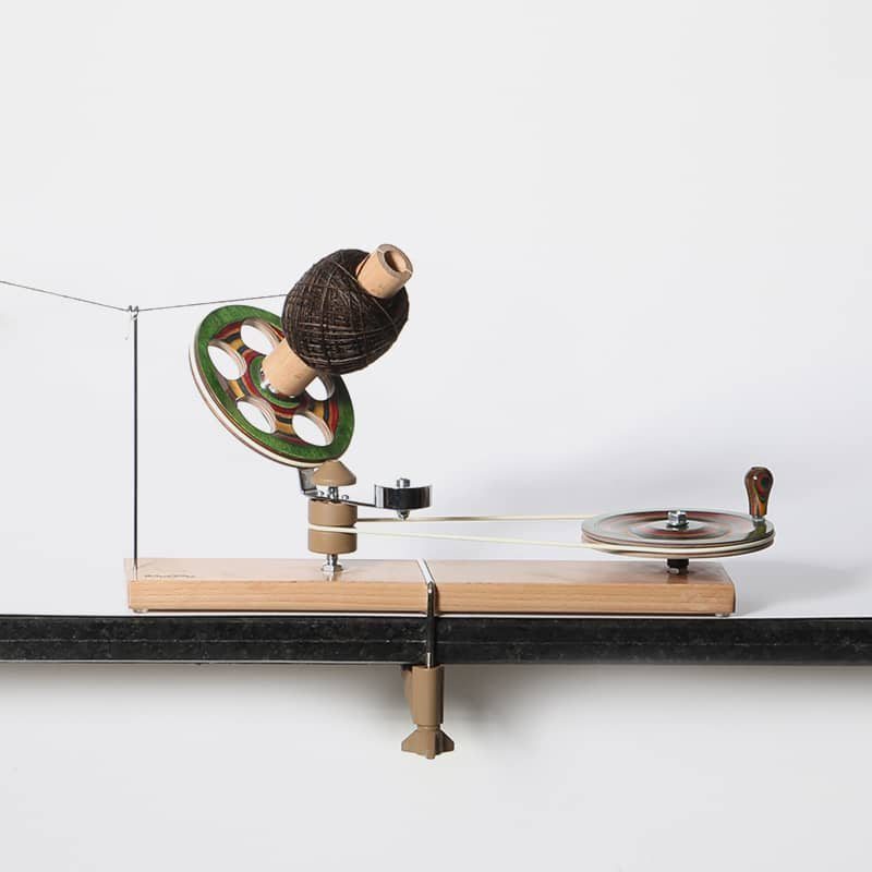 A tabletop hand-crank yarn winder is secured to a wooden surface with a clamp. The device has a brown yarn spool, a green circular component with three holes, and a wooden handle for manual operation. A thin wire is connected to the setup.